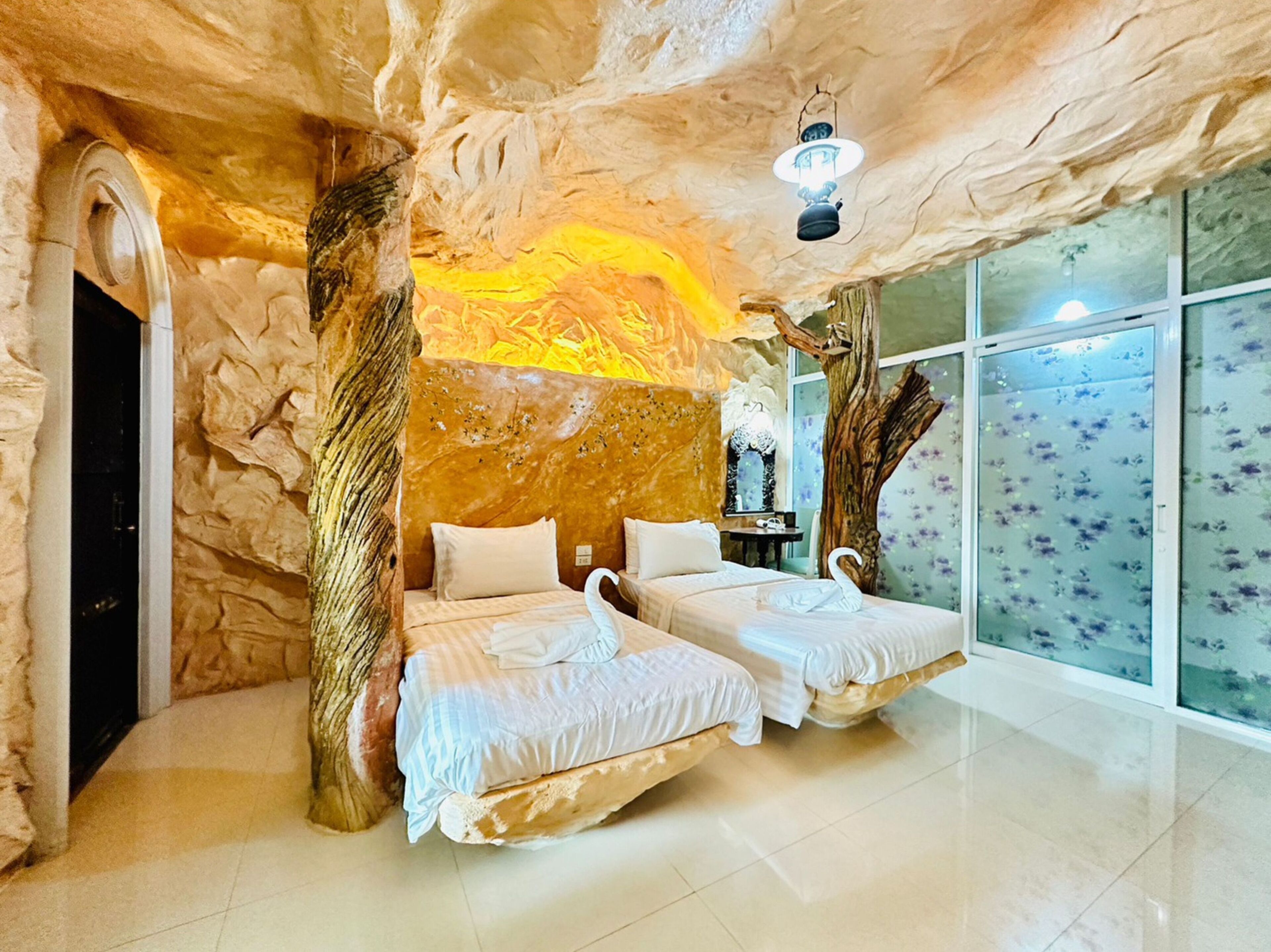 A room with two beds, a large window with floral blinds, and a unique rock-like ceiling.