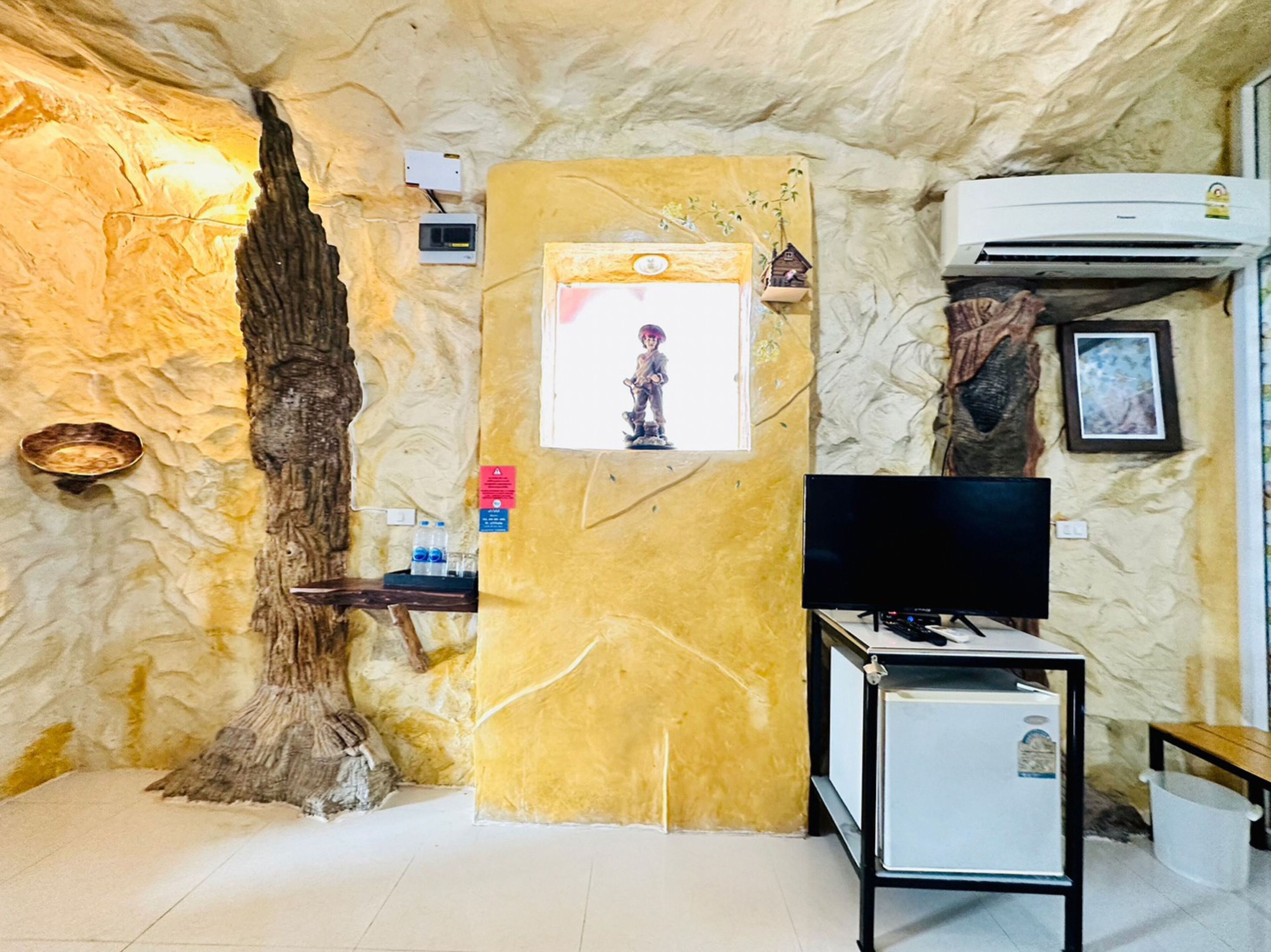 Una habitación con pared de piedra, una escultura de árbol de madera, un televisor, un refrigerador pequeño y un equipo de aire acondicionado.