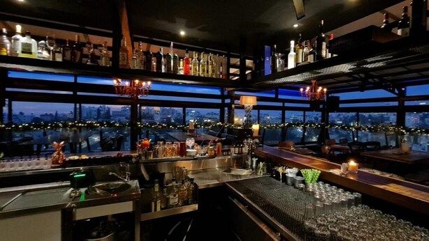 A bar area with multiple shelves of bottles, a bar counter, and a view of the cityscape through large windows. - Ankars Suites & Hotel (Ramallah)