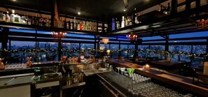 Zona de bar con varias estanterías llenas de botellas, una barra y vistas panorámicas de la ciudad a través de amplios ventanales.
