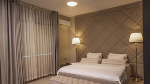 A hotel room with a large bed, two bedside lamps, a tufted bench, and a window with curtains. - Ankars Suites & Hotel (Ramallah)
