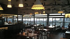 A restaurant with high ceilings, hanging pendant lights, and large windows offering city views. Tables and chairs are arranged for dining. - Ankars Suites & Hotel (Ramallah)