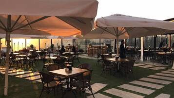 An outdoor dining area with tables and chairs, covered by large umbrellas, and a view of the cityscape through large windows.