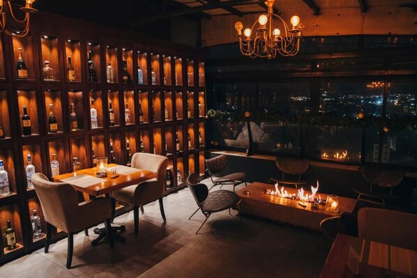 Zona de bar con una larga mesa de madera, sillas acolchadas, una pared con botellas de licores y una chimenea.