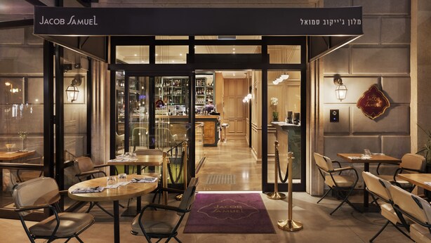 Un restaurant avec des tables et des chaises disposées à l’extérieur, une enseigne indiquant « Jacob Samuel », et un intérieur visible avec des étagères et un comptoir.