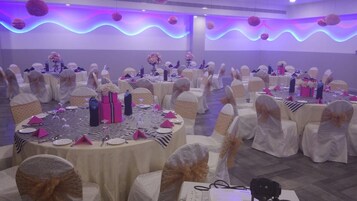 Un salón de banquetes con mesas redondas dispuestas para un evento formal, con sillas blancas adornadas con lazos decorativos, mesas cubiertas con manteles a rayas y centros de mesa florales.