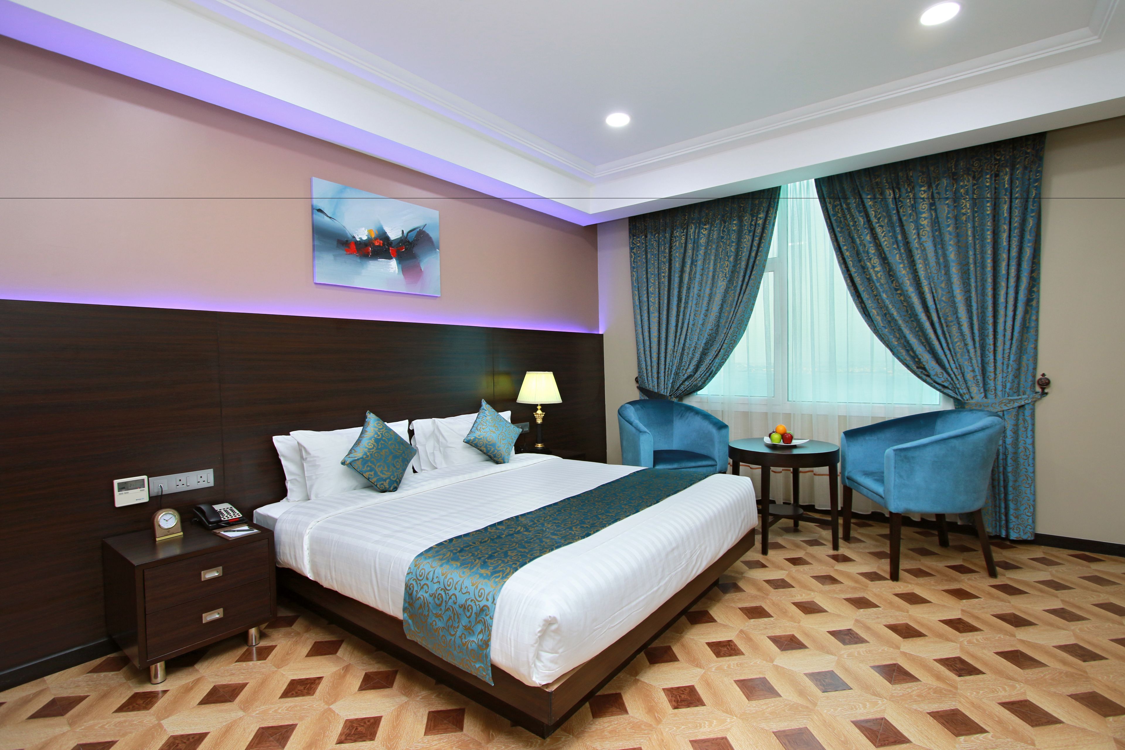 Quarto de hotel com uma cama grande, mesa de cabeceira com abajur, pequena mesa com frutas e duas cadeiras.