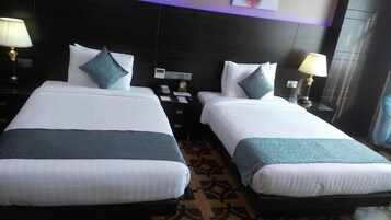 Habitación de hotel con dos camas, cada una con edredón blanco y almohada decorativa azul. Se observan mesitas de noche con lámparas y un reloj sobre una de ellas.