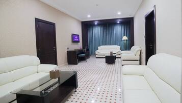 Habitación de hotel moderna con dos sofás blancos, una mesa de centro de vidrio, un televisor de pantalla plana y una puerta de madera oscura.