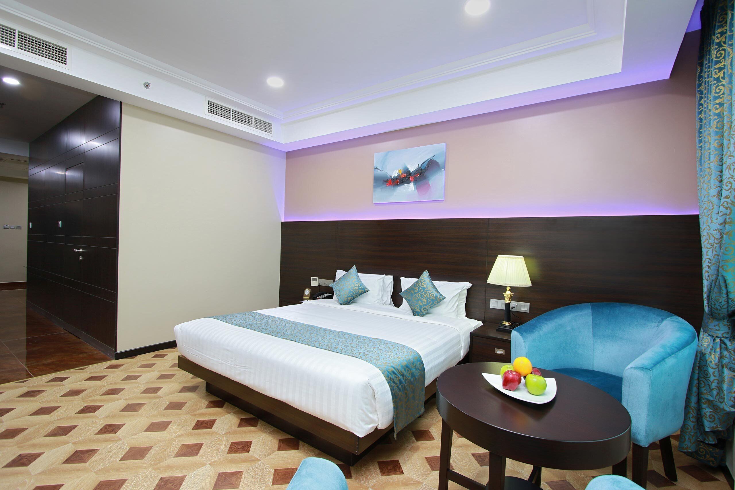 A hotel room with a large bed, a bedside table with a lamp, a blue armchair, and a fruit bowl.