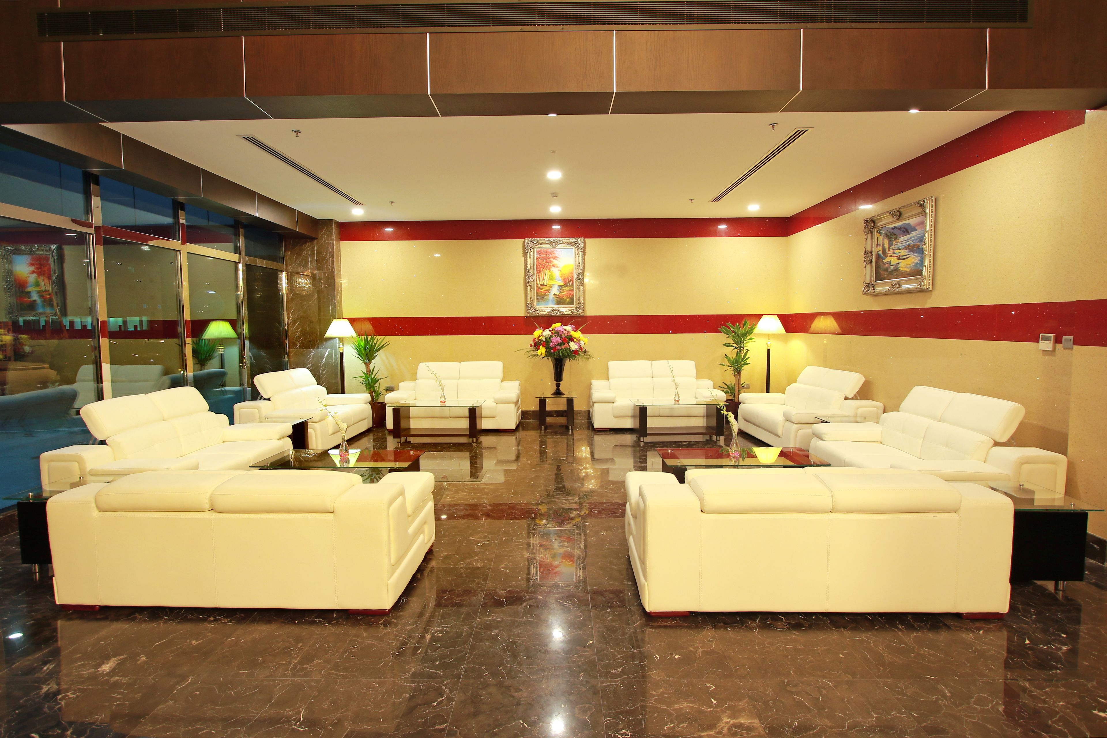 Un hall con sofás de cuero blanco dispuestos en forma de U, suelo de mármol pulido y una mesa central con un jarrón de flores.