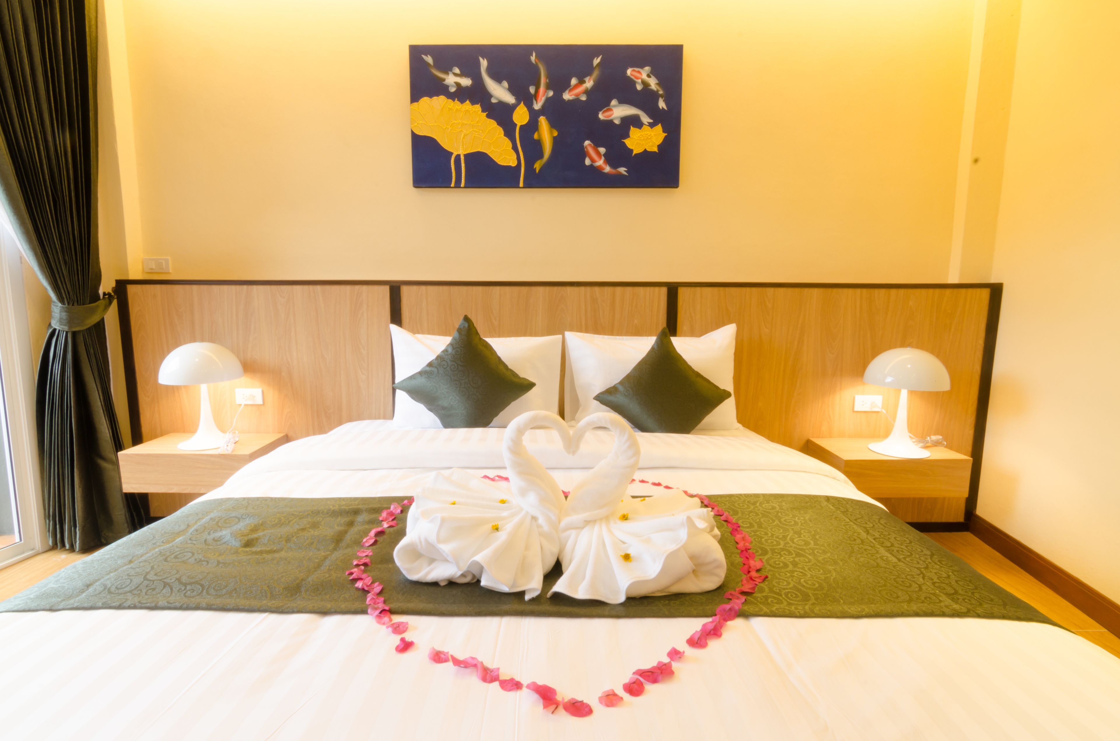 A hotel room with a neatly made bed, two bedside lamps, a painting on the wall, and a heart-shaped decoration on the bed.