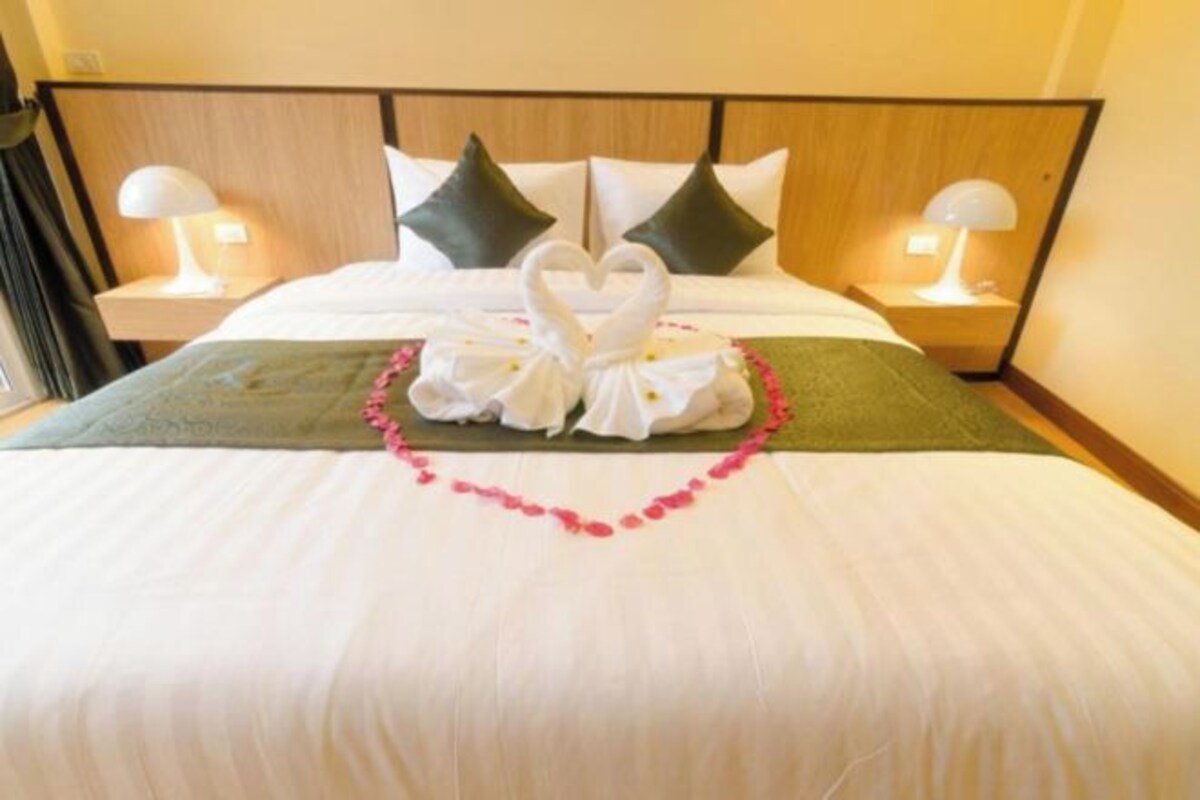 A hotel room with a neatly made bed, two pillows, a green throw blanket, and a heart-shaped towel arrangement on the bed.