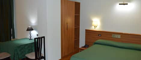 Habitación de hotel con cama, escritorio con lámpara, silla, armario y mesita de noche con lámpara.