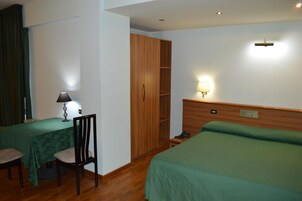 Habitación de hotel con cama, escritorio con lámpara, silla, armario y mesita de noche con lámpara.