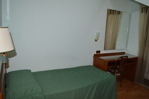 A small, single-bed room with a green bedspread, a wooden desk, a chair, a lamp, and a mirror on the wall.