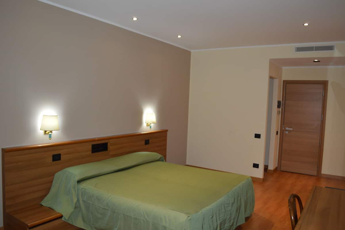 A hotel room with a single bed, two bedside lamps, a wooden headboard, a wooden table, and a door.