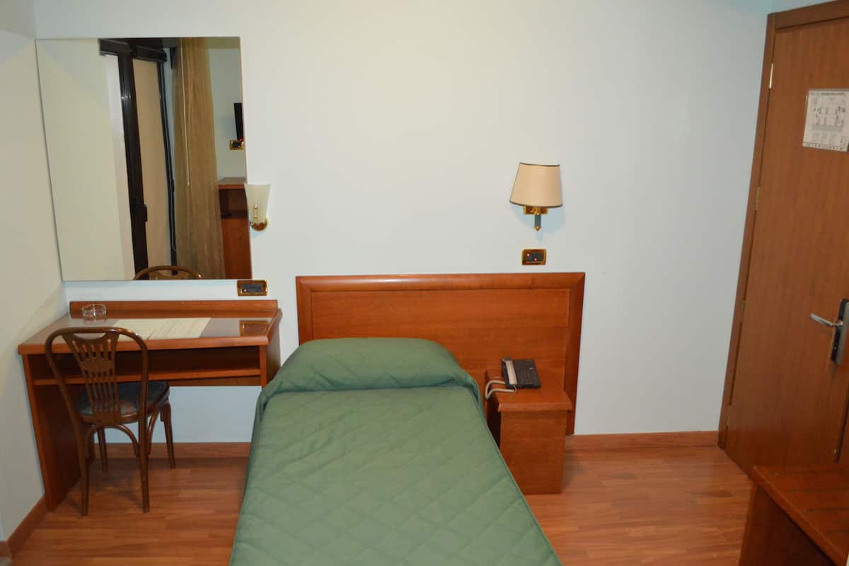 A hotel room with a wooden desk, a chair, a bed with a green bedspread, a bedside table with a lamp, and a door with a lock.