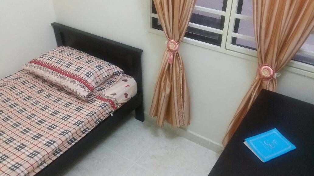 A small, single-bed room with a checkered bedspread, a nightstand with a blue card, and curtains tied back to reveal a window.