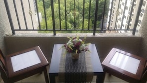 Un balcon avec une table et deux chaises, un arrangement floral en guise de décoration centrale, et une vue sur la verdure et les bâtiments.