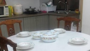 Une table de salle à manger ronde, entourée de six chaises, et complétée par des meubles en bois et un coin cuisine équipé d’appareils électroménagers et de vaisselle.