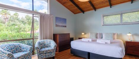 A bedroom with a large window, a bed, two patterned armchairs, a dresser, and a nightstand with lamps.