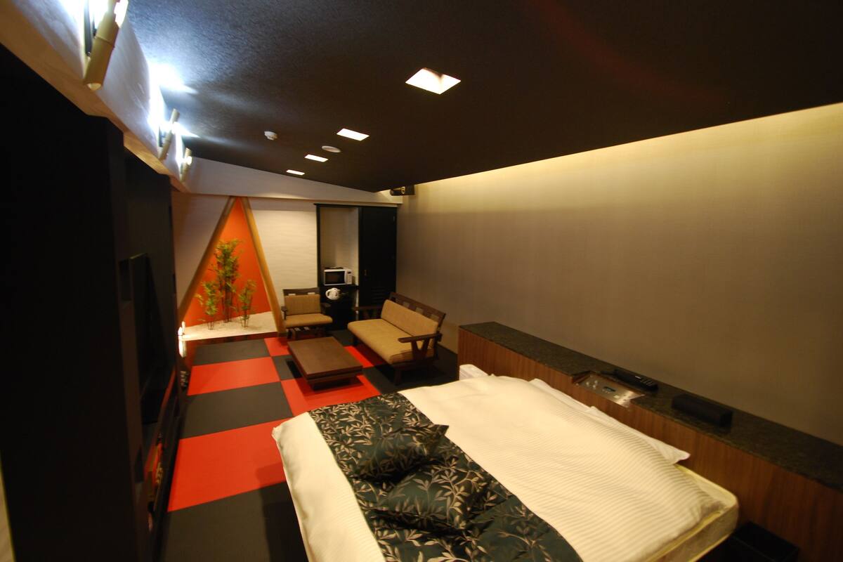 A hotel room with a bed, a desk, a chair, a small table, and a plant.