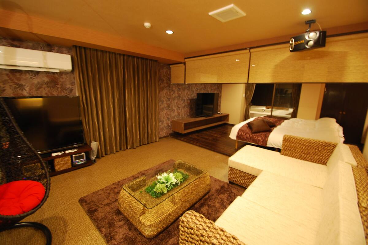 A hotel room with a flat-screen TV, a sofa, a small table with a decorative item, a wicker chair, and a bed with a white comforter.