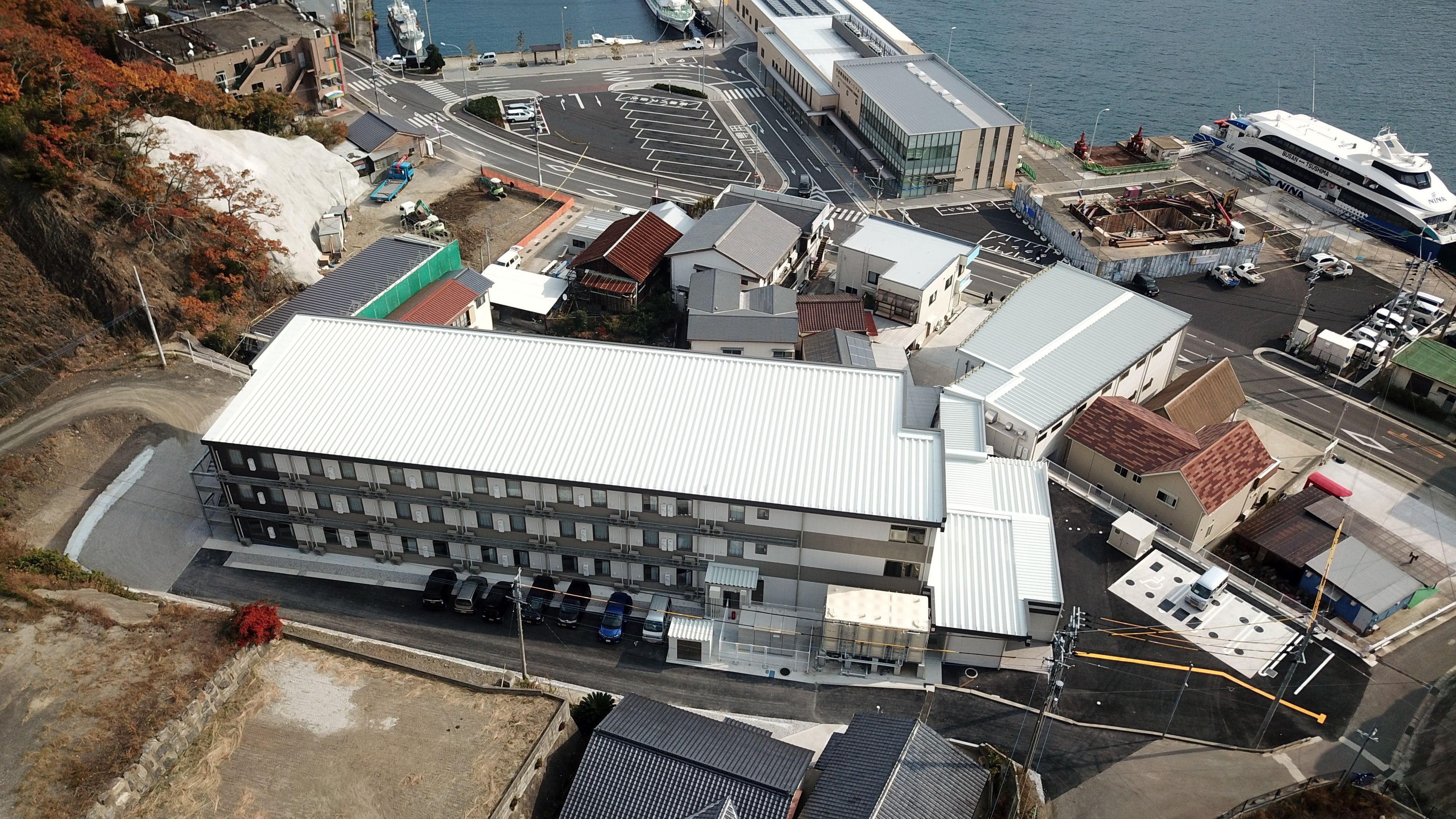 An aerial view of a coastal area with a large building, a marina with boats, and a road leading to the marina.