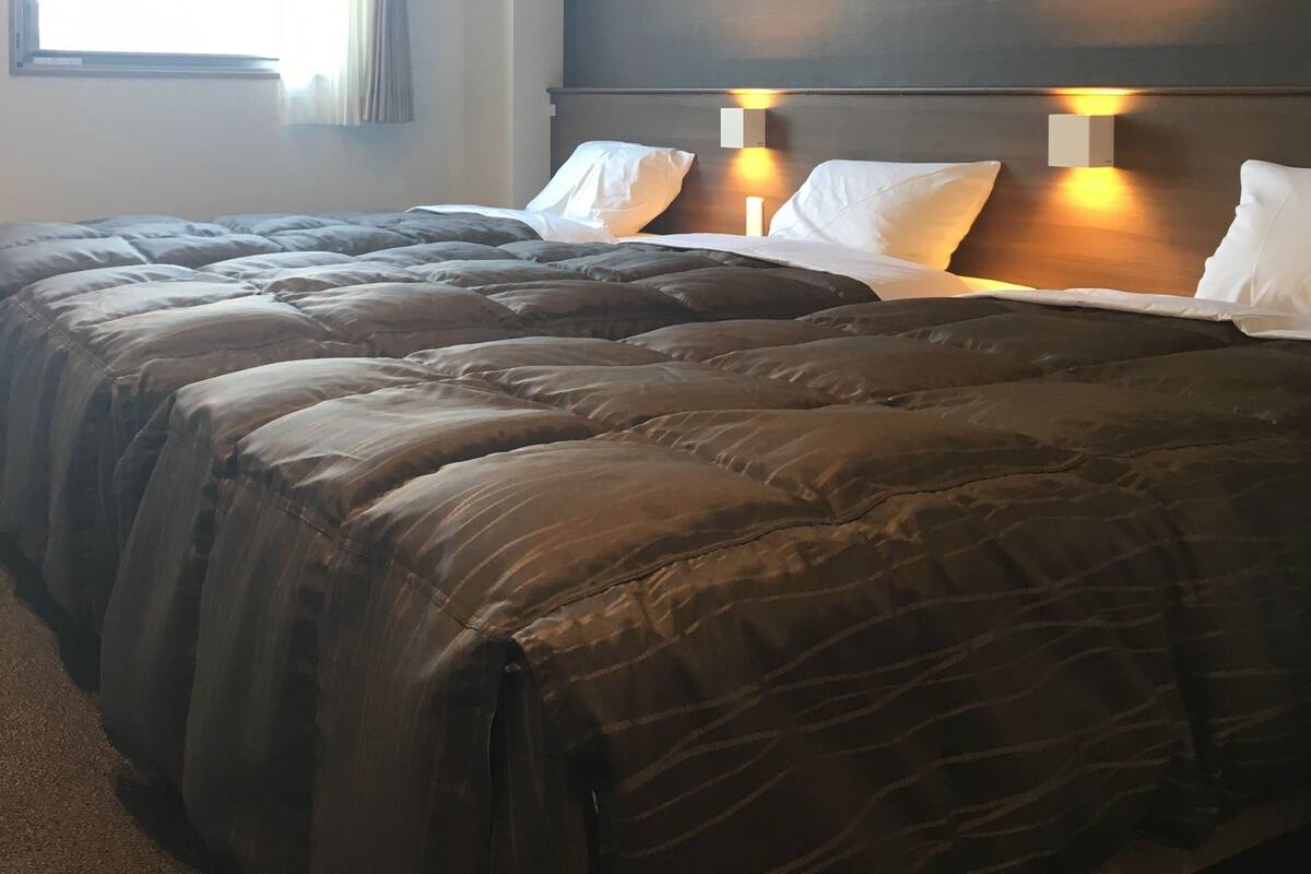 A hotel room with a large bed, two bedside lamps, a window with curtains, and a plain wall.