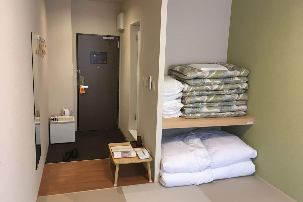 A hotel room with a wooden floor, a small table with books, a bed with white bedding, a shelf with folded blankets, and a door with a sign.