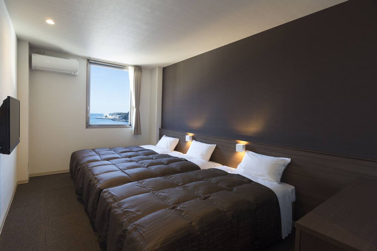 A hotel room with two beds, a large window, an air conditioning unit, and a view of the sea.