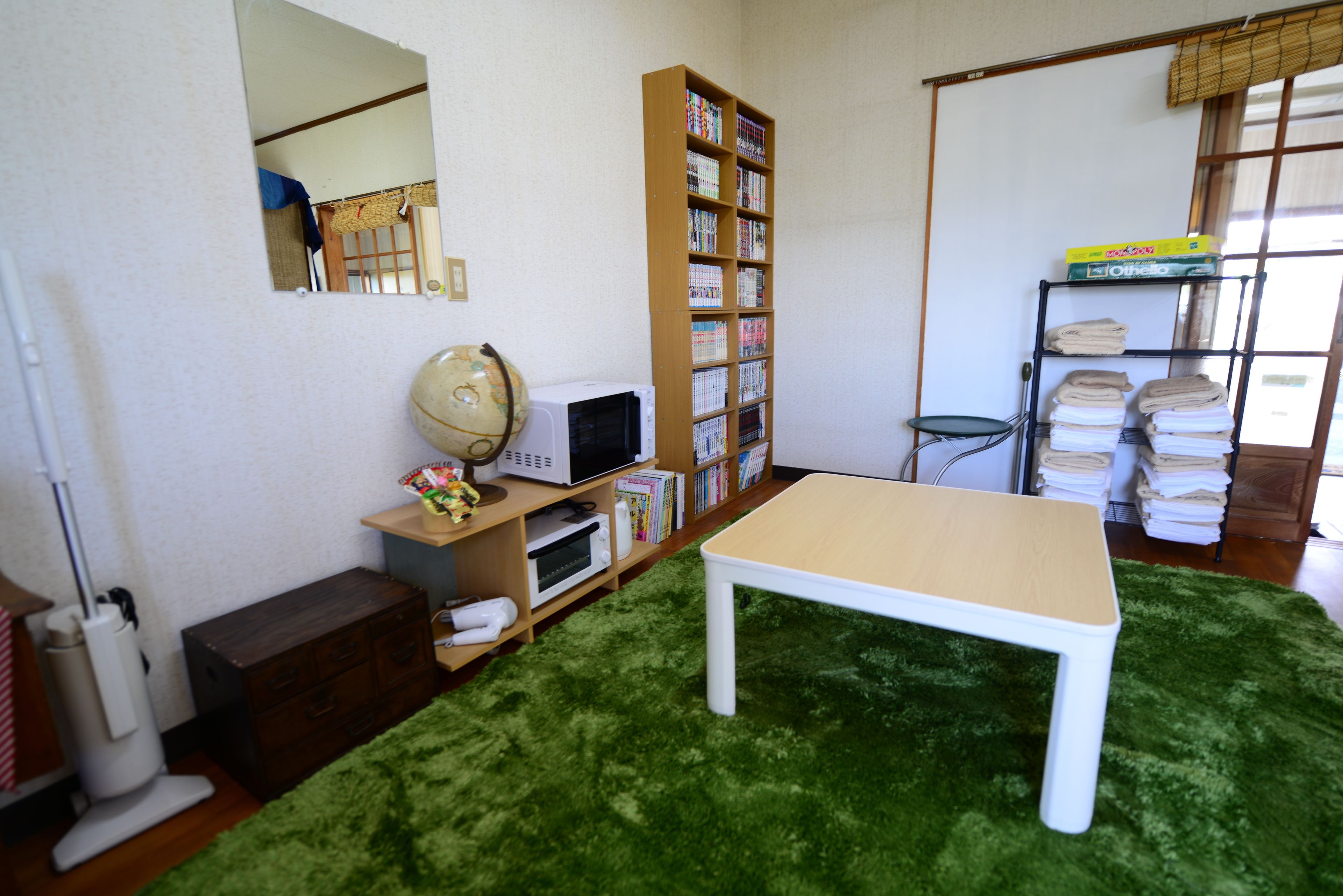 A room with a green carpet, a white table, a bookshelf, a mirror, and a window with blinds.