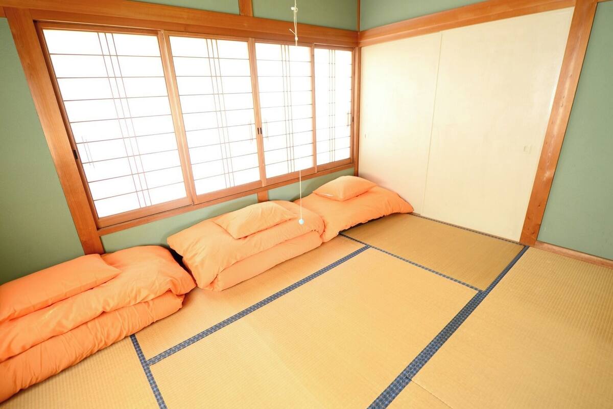 Ein Raum mit Tatami-Matten, einem Futonbett, Schiebetüren und Blick durch ein Fenster mit Shoji-Bildschirmen.