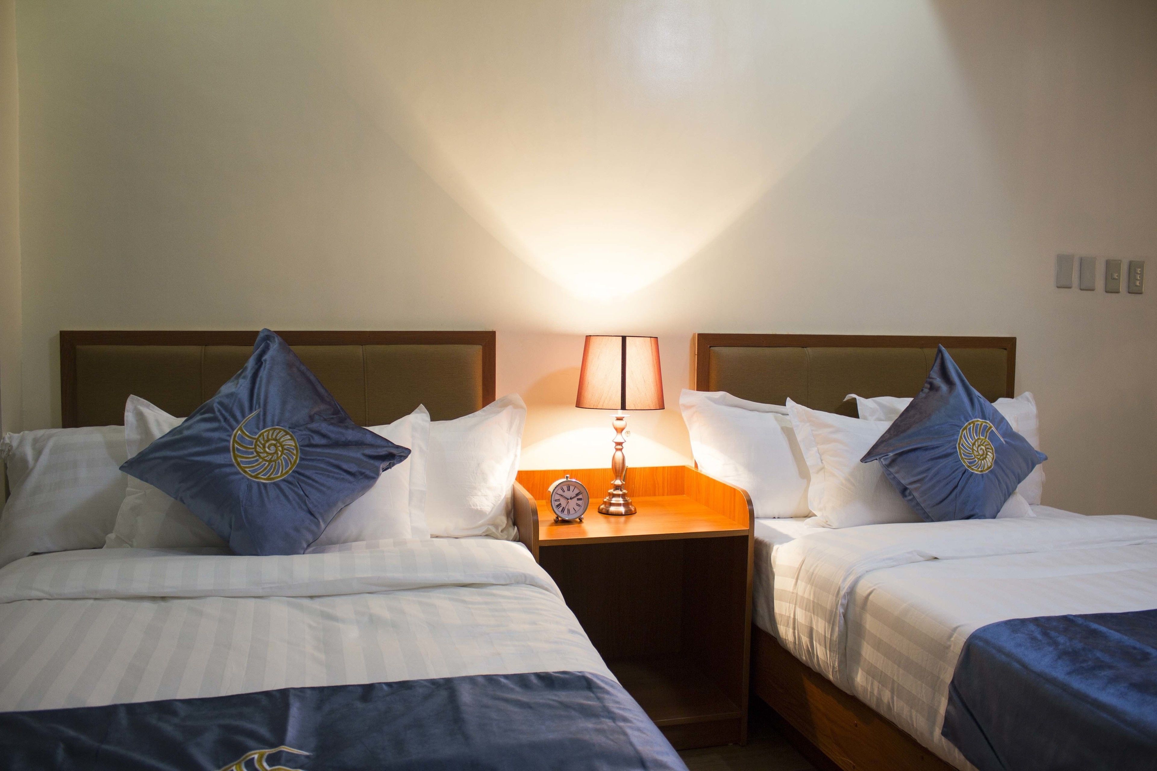 Une chambre d’hôtel avec deux lits, chacun agrémenté d’un oreiller bleu et d’un oreiller blanc. Une table de chevet avec une lampe et une horloge se trouve entre les deux lits.