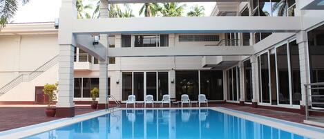 A swimming pool with white chairs and potted plants, surrounded by a building with large windows and a covered area.