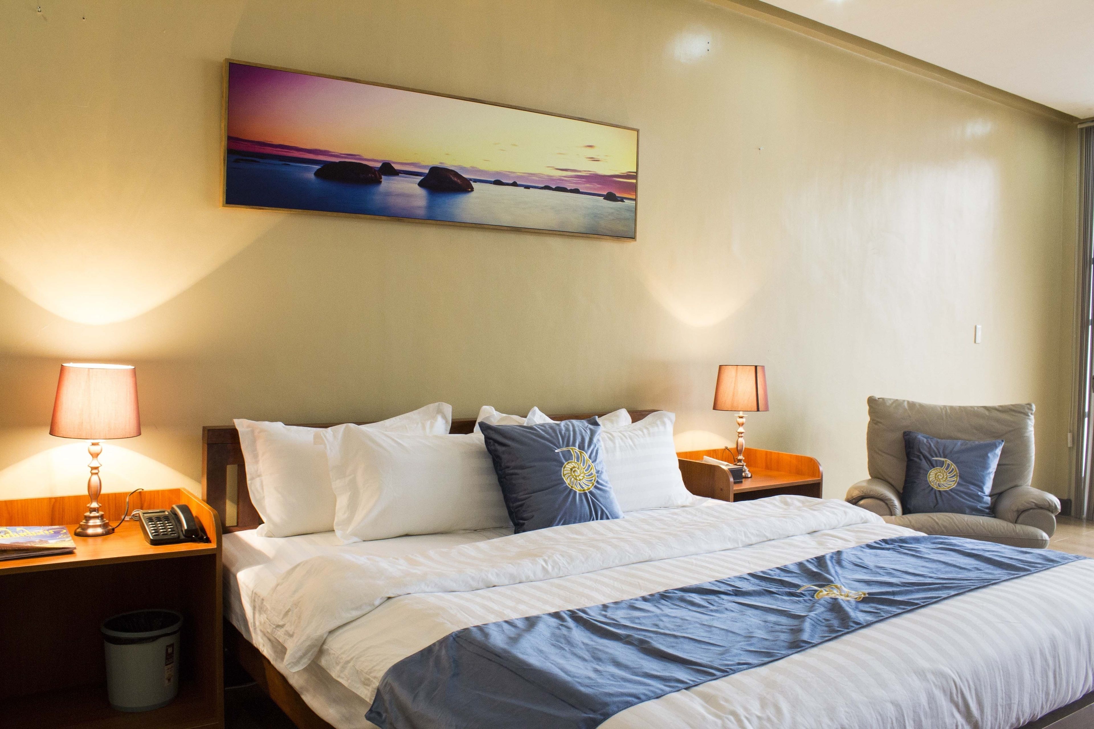 A hotel room with a bed, two bedside tables, a chair, a painting on the wall, and a nightstand with a lamp and phone.