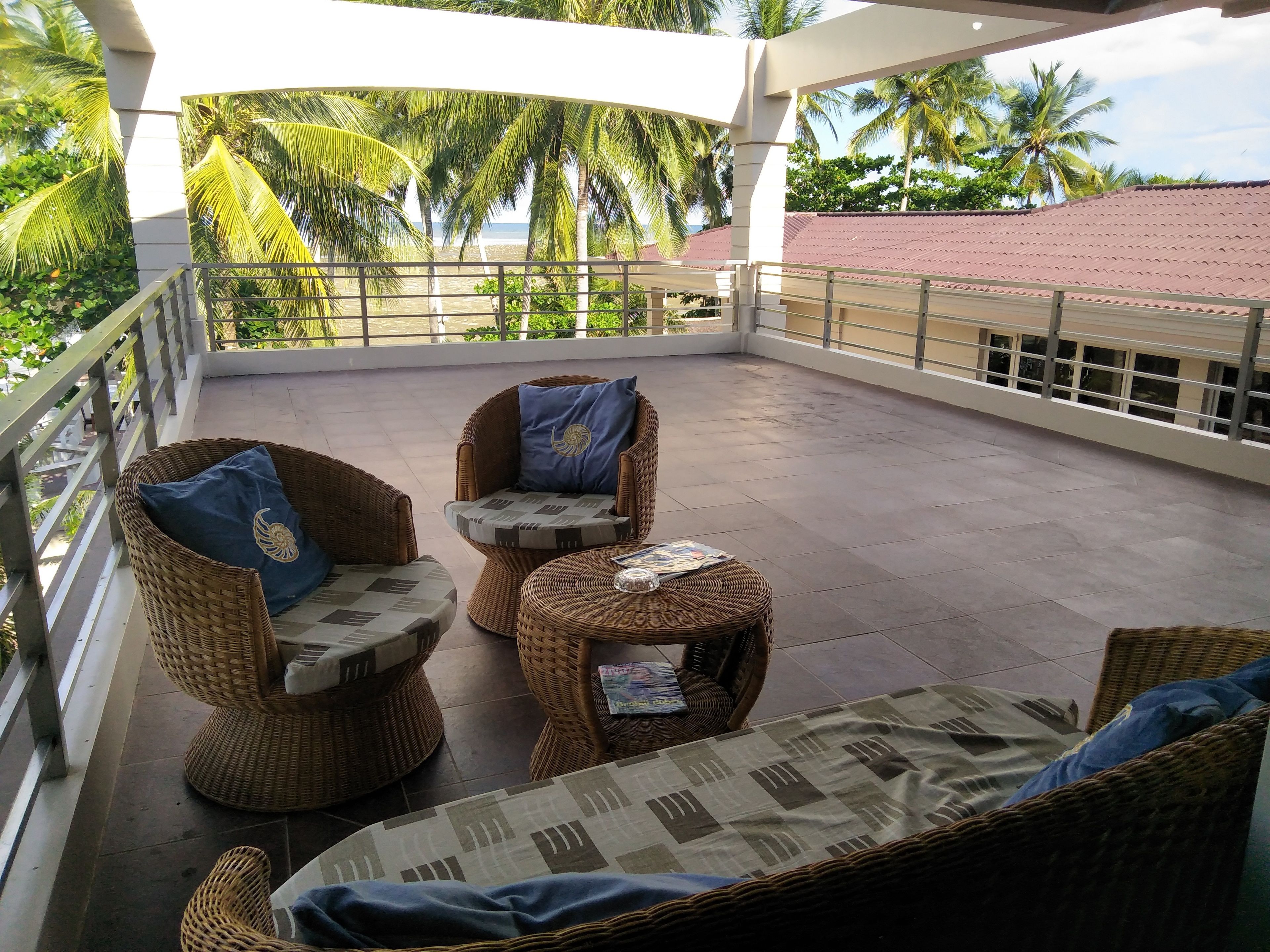 A covered balcony with wicker furniture, a tiled floor, and a view of palm trees and a beach.