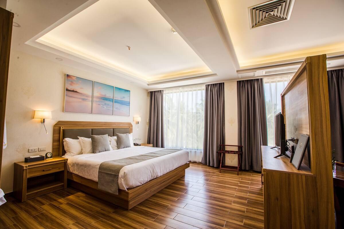 A modern hotel room with a large bed, wooden flooring, a desk with a computer, and a large window with curtains.