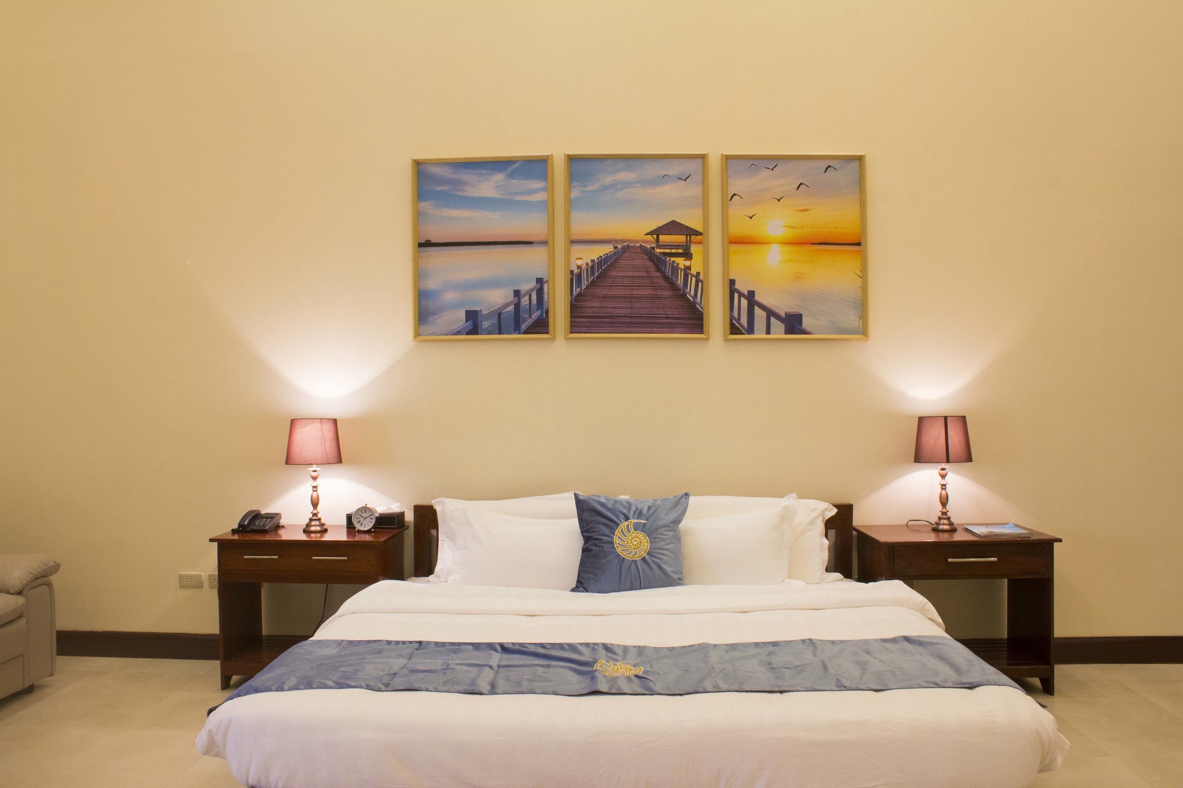 Une chambre d’hôtel avec un lit, deux tables de chevet, une lampe, une horloge et trois tableaux accrochés au mur.