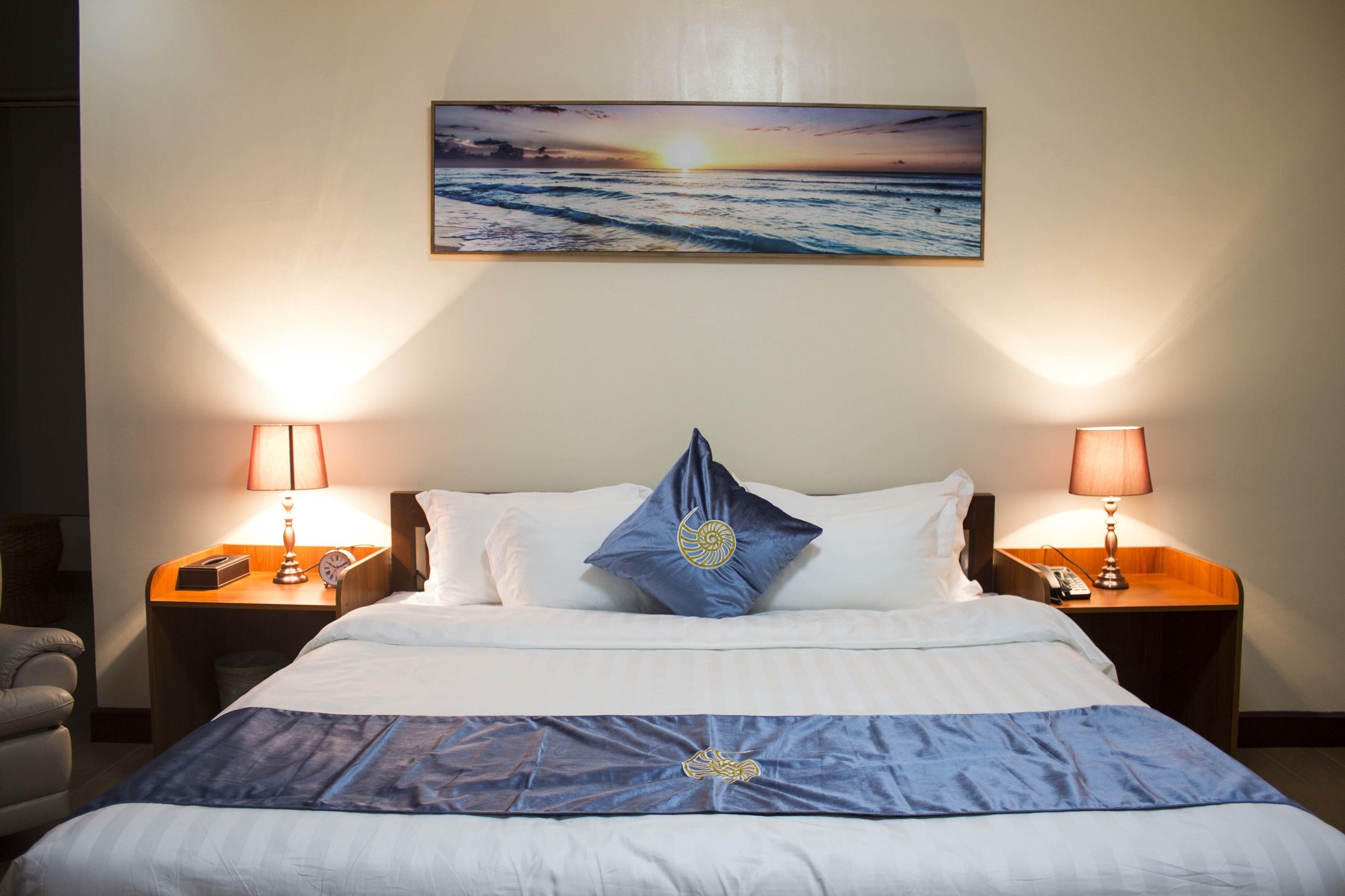 A neatly made bed with a blue and white bedspread, two bedside lamps, a nightstand with a phone, and a painting of a sunset over the ocean on the wall.
