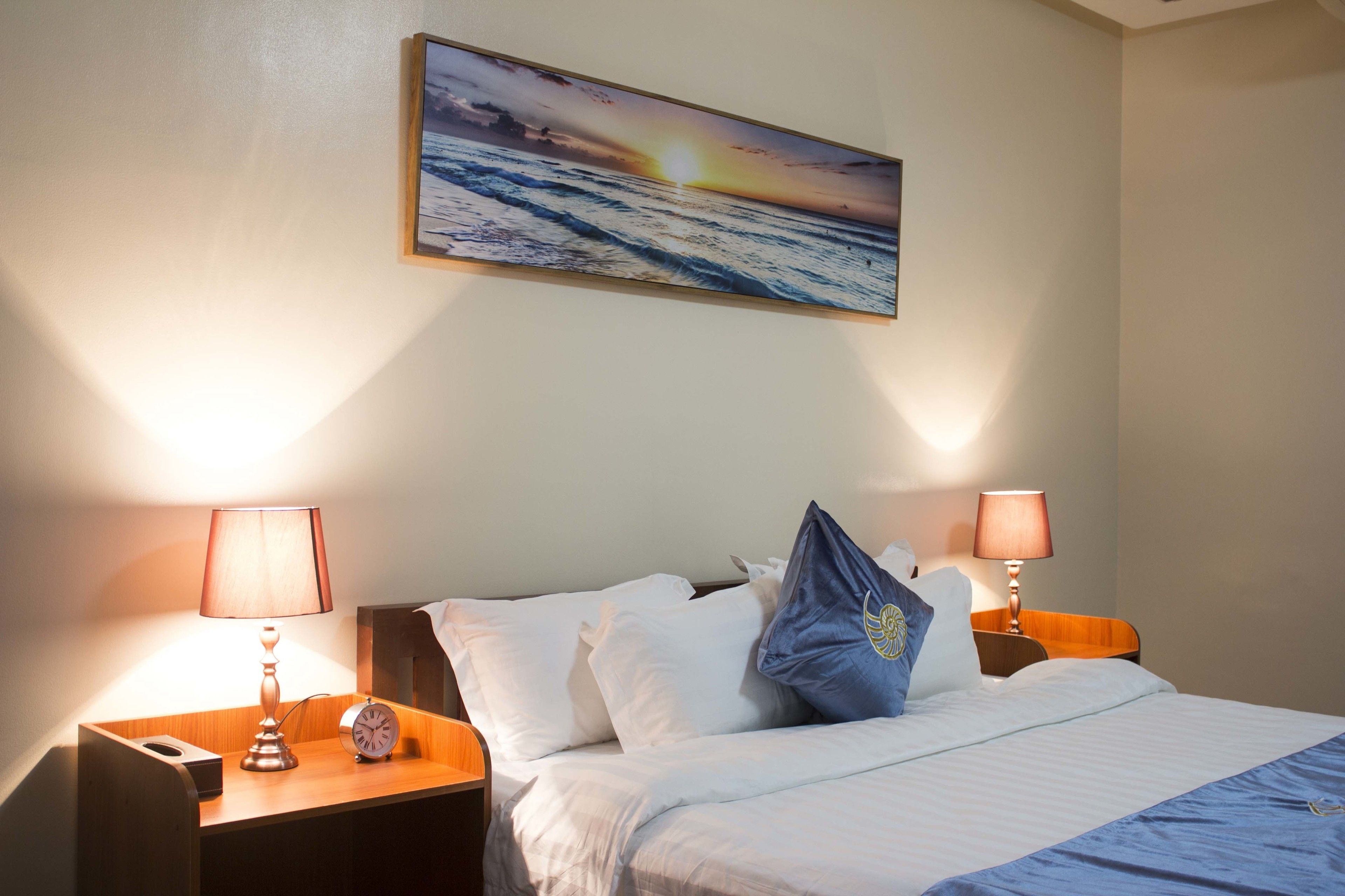 A hotel room with a bed, two bedside tables, lamps, a clock, and a painting of a sunset over the ocean.