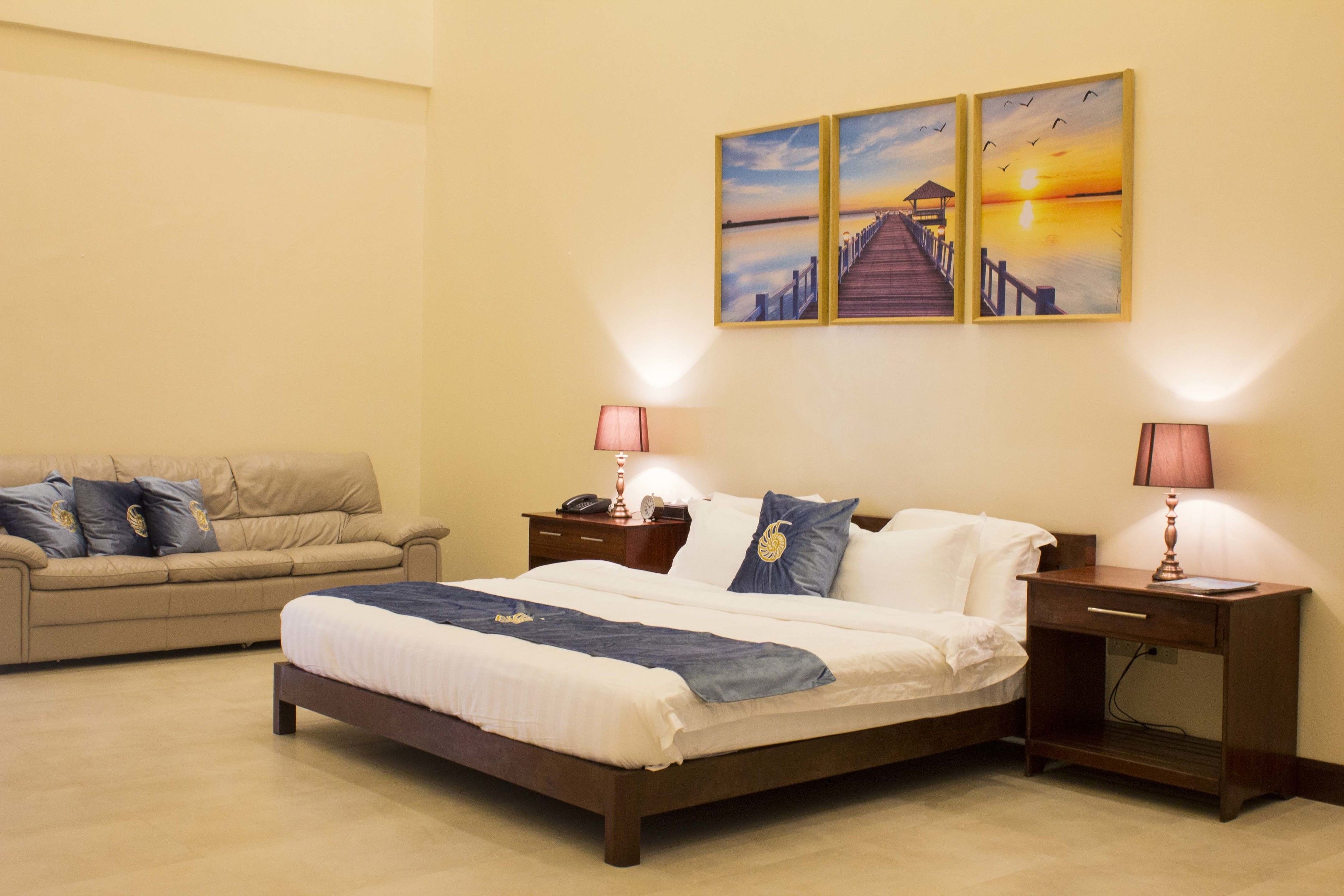A hotel room with a bed, a nightstand, a lamp, a sofa, and a wall with three paintings.