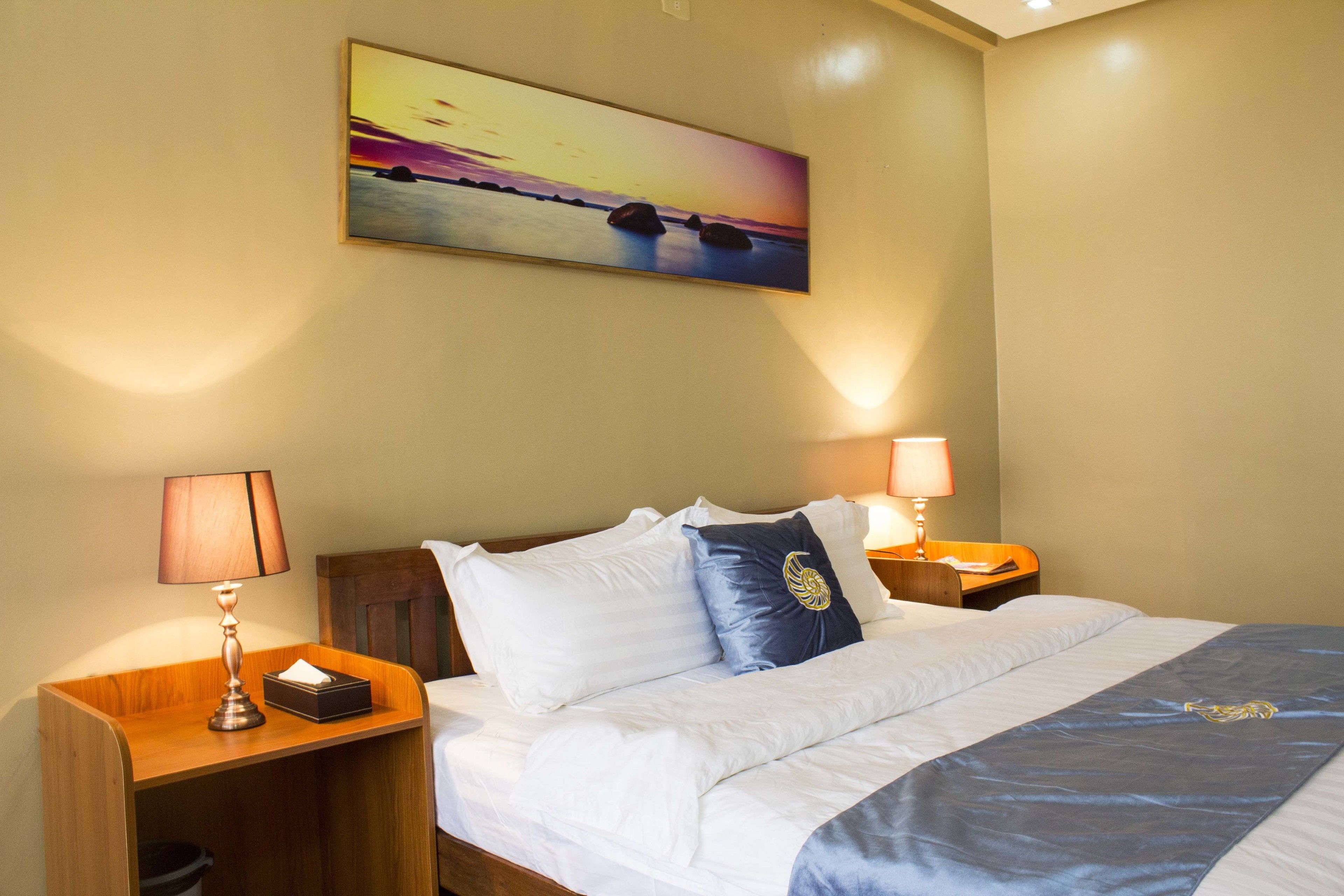 Habitación de hotel con cama, lámparas de noche, mesita de noche, un cuadro de un atardecer y un cabecero de madera.