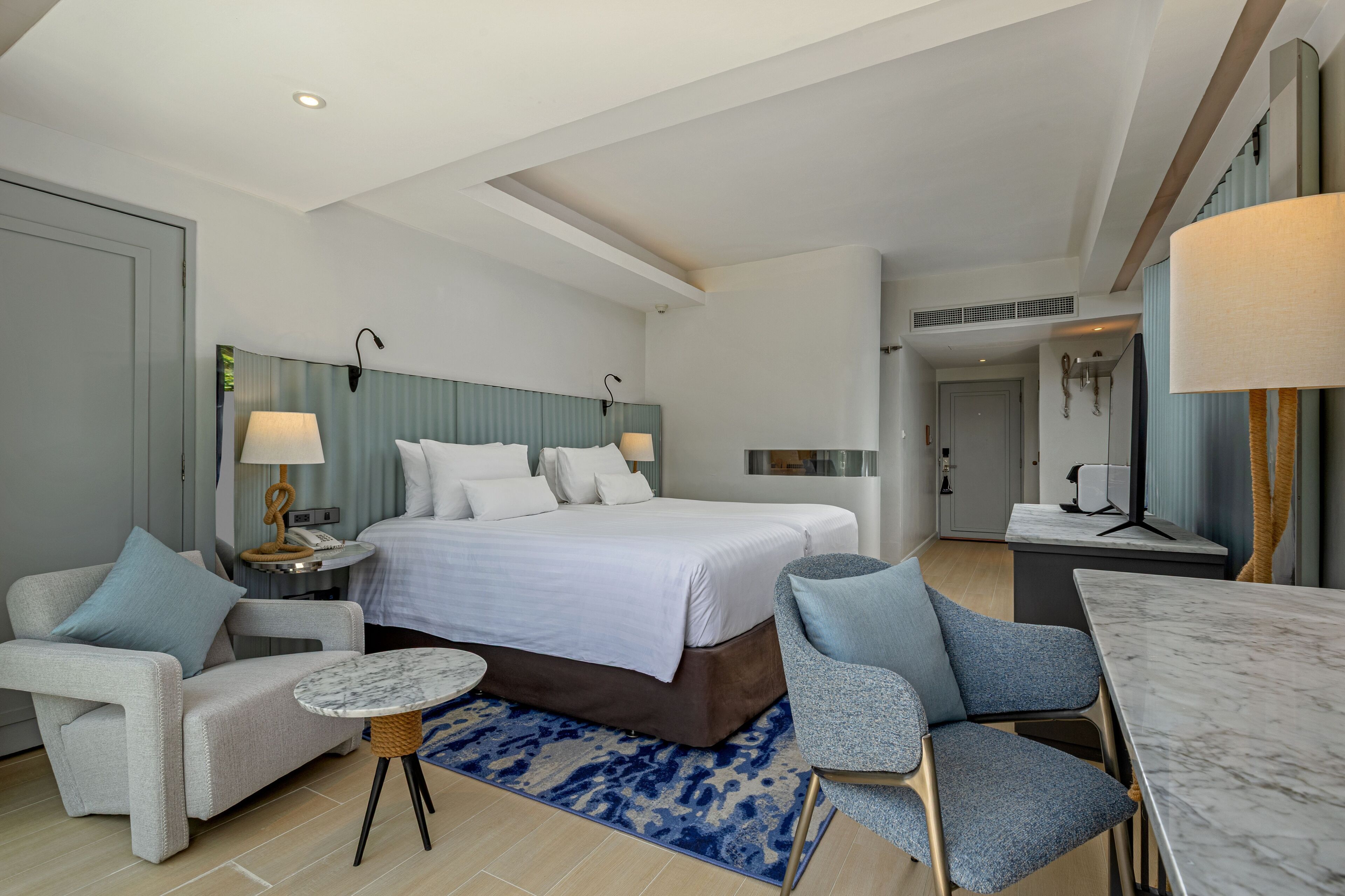 Habitación de hotel con una cama grande, dos sillones, una mesita redonda, un escritorio y un baño visible a través de una puerta.