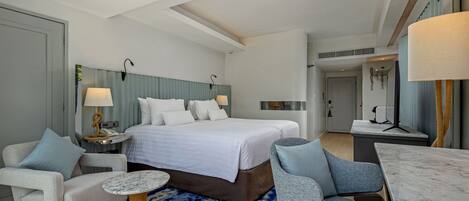 Habitación de hotel con una cama grande, dos sillones, una mesita redonda, un escritorio y un baño visible a través de una puerta.