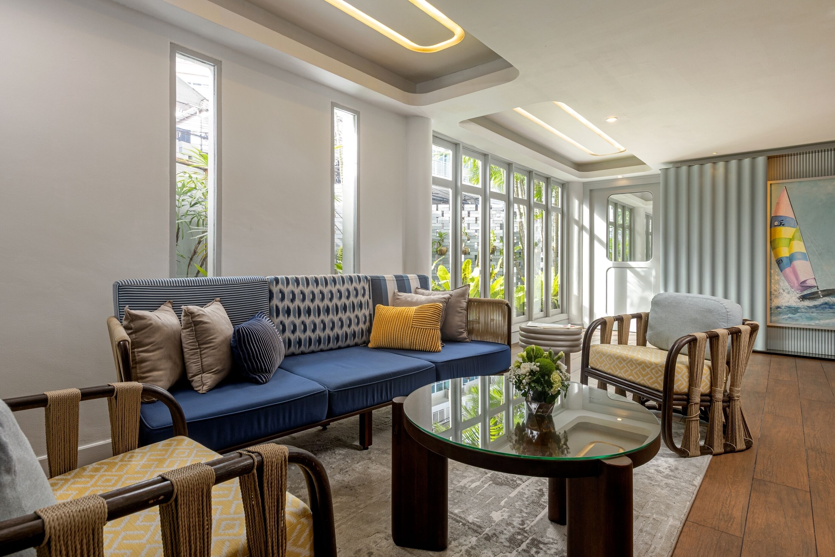 A modern living room with a blue sofa, wicker chairs, a glass coffee table, and large windows with a view of greenery.