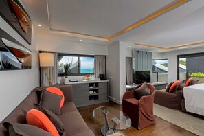 Una habitación de hotel moderna con sofá, una mesa de centro de vidrio, televisión, cama y vistas al mar.