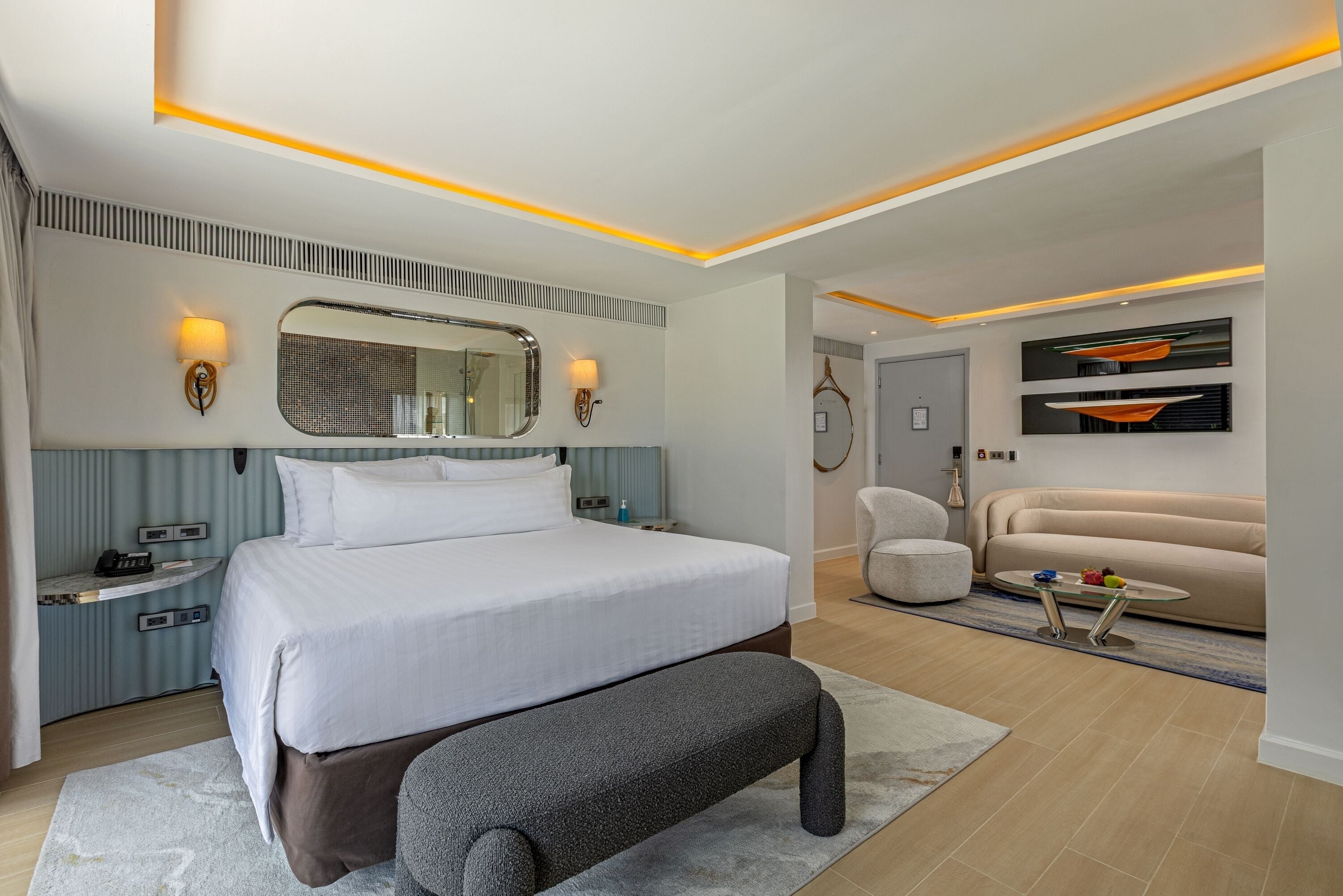 A modern hotel room with a large bed, bedside tables, a bench, a seating area with a sofa and armchairs, and a small table with a tray.