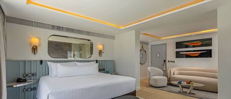 A modern hotel room with a large bed, bedside tables, a bench, a seating area with a sofa and armchairs, and a small table with a tray.